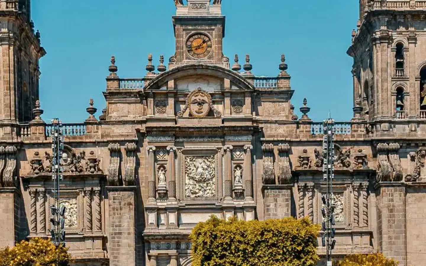 A wide shot of the Metropolitan Cathedral of Mexico City, a grand Baroque structure with two bell towers, a central clock, and intricate stone carvings, under a clear blue sky.