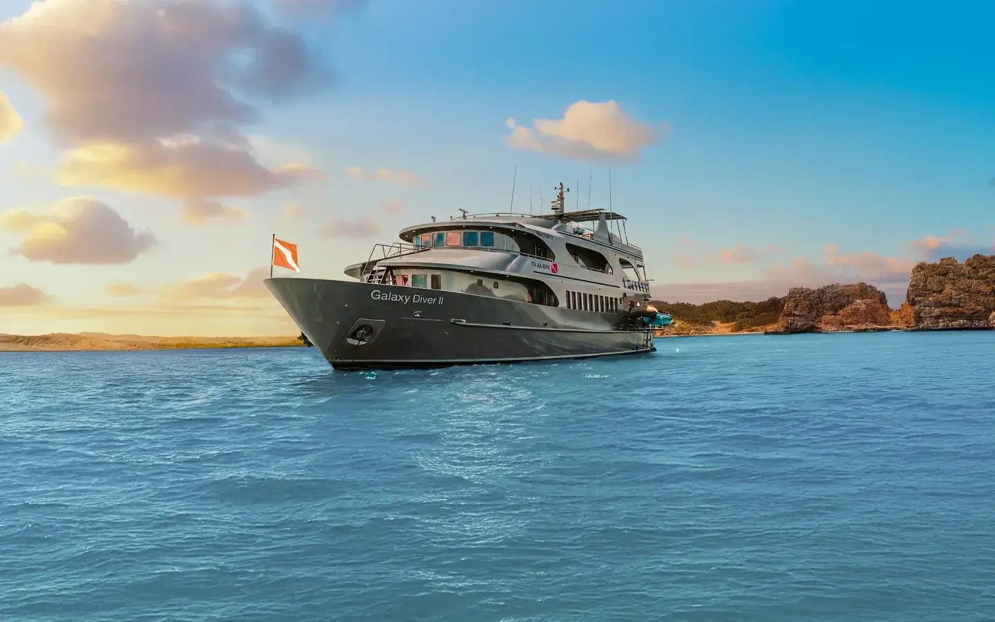 Galaxy Diver II at anchor at sunset off a Galápagos coastline, hull name visible on the bow