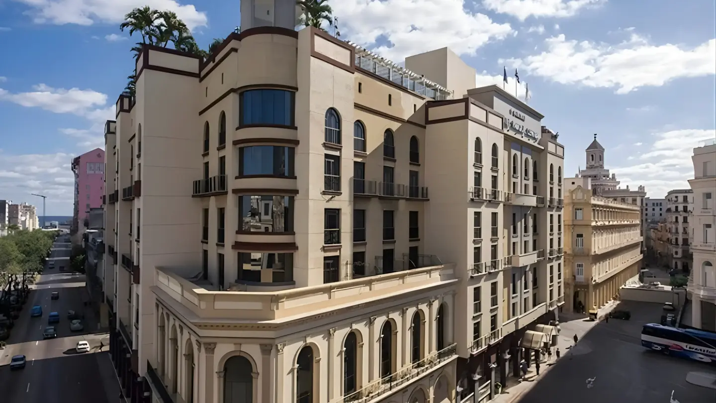 An aerial view of a cream-colored hotel with a domed tower, palm trees on its roof, and arched windows, situated on a street with cars and a blue bus in a sunny, cloudy urban environment.