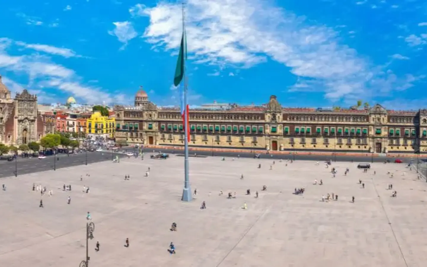 A panoramic view of the Zócalo in Mexico City, featuring the Metropolitan Cathedral, the National Palace, and a large flagpole with the Mexican flag under a bright blue sky.