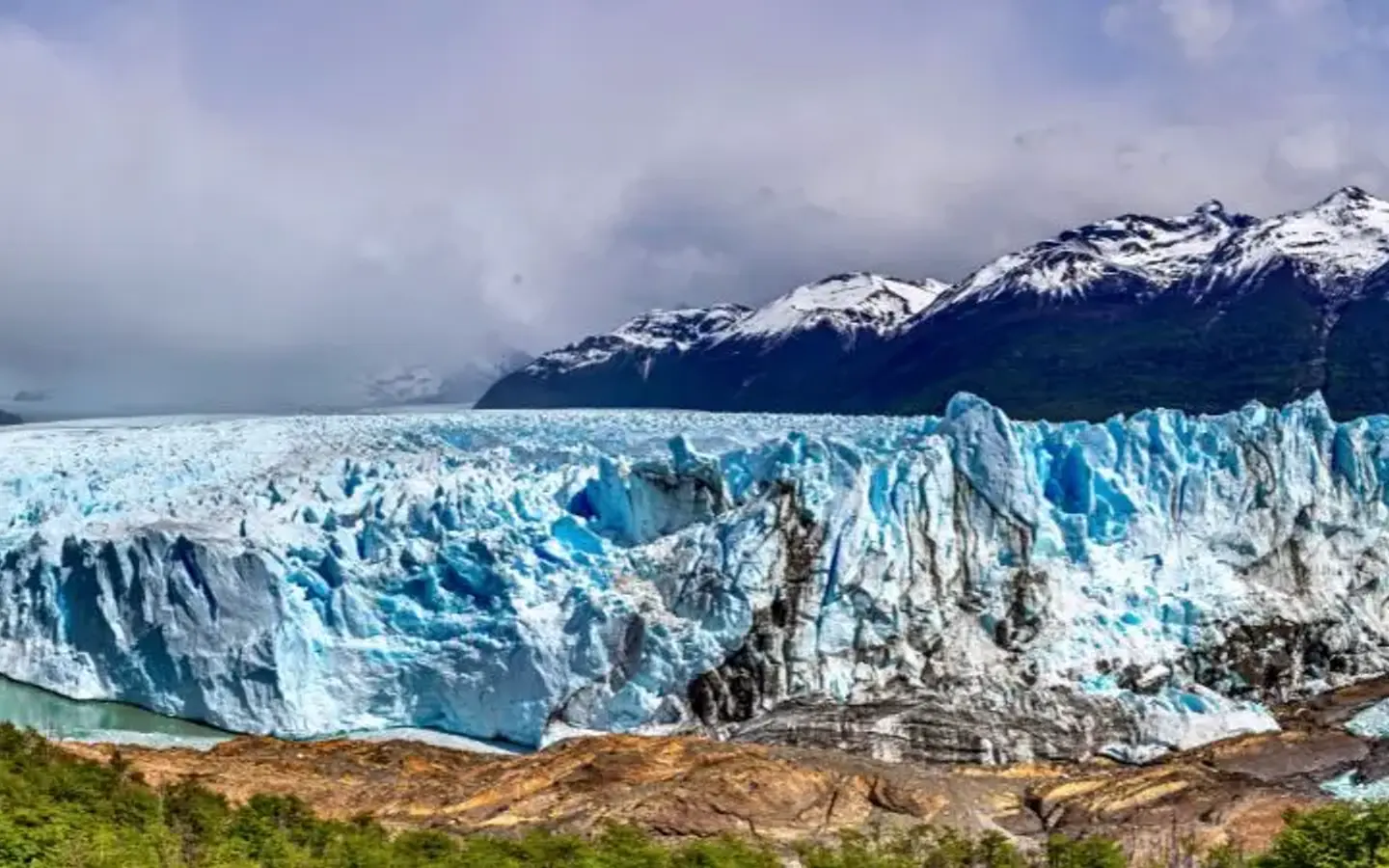 A vast, blue-tinged glacier juts into a turquoise lake, backed by snow-capped mountains and lush green hills under a cloudy sky.