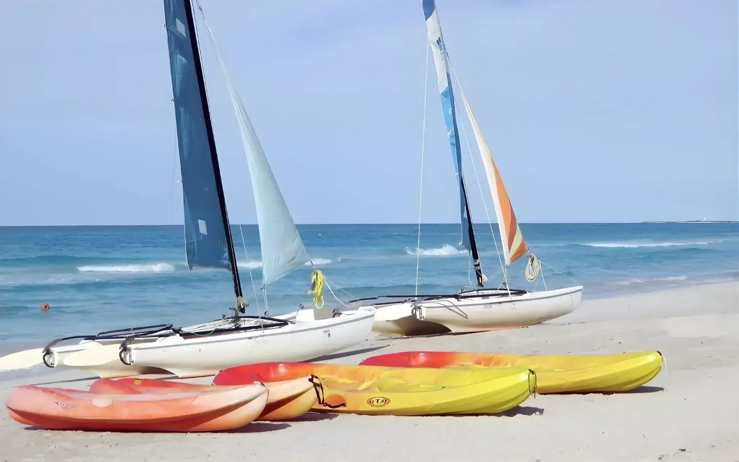 Two white catamarans with sails are parked on a sandy beach, with three colorful kayaks in the foreground and the blue ocean and sky in the background.