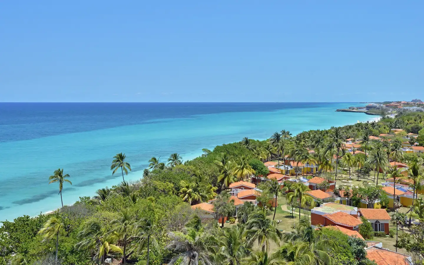 A panoramic view of a tropical resort with orange-roofed buildings nestled among lush palm trees and vibrant green foliage, overlooking a stunning turquoise ocean under a clear blue sky.